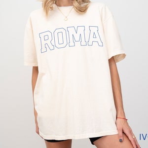 May include: A white oversized t-shirt with a blue outline of the word "ROMA" printed on the front. The shirt is being modeled by a person. The background is white.