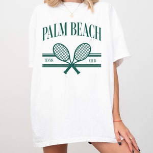 May include: White t-shirt with a green graphic of two crossed tennis rackets and the text "PALM BEACH TENNIS CLUB".