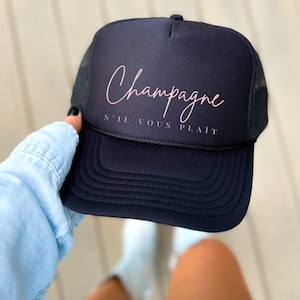 May include: Black trucker hat with a pink cursive script that says "Champagne S'il Vous Plaît"