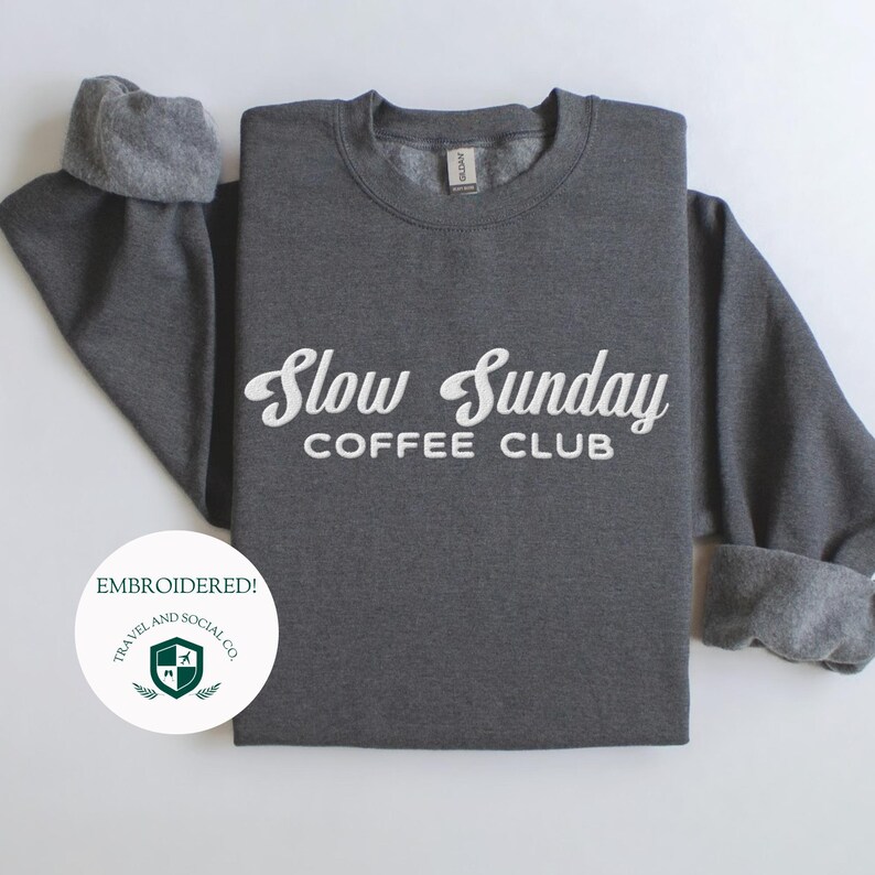 May include: Gray sweatshirt with the text "Slow Sunday Coffee Club" embroidered in white. The sweatshirt is folded and displayed on a white background. A small green logo with the text "Travel and Social Co." is in the lower left corner.