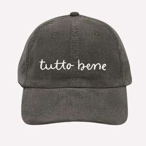 May include: A dark olive green corduroy baseball cap with the white cursive text "tutto bene" embroidered on the front. The cap has a curved brim and a fabric-covered button on top.