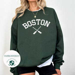 Puede incluir: Una sudadera verde oscuro con la palabra "BOSTON" bordada en blanco con dos remos cruzados debajo. La sudadera está hecha de un material suave y cómodo y tiene un corte holgado.