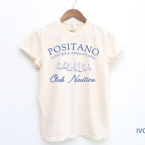 May include: An ivory short-sleeve t-shirt featuring blue text that reads POSITANO COSTIERA AMALFITANA and Club Nautico, with a graphic illustration of several sailboats positioned between the two lines of text, hanging on a wooden hanger.