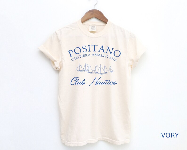 May include: An ivory short-sleeve t-shirt featuring blue text that reads POSITANO COSTIERA AMALFITANA and Club Nautico, with a graphic illustration of several sailboats positioned between the two lines of text, hanging on a wooden hanger.