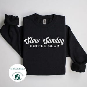 May include: Black sweatshirt with white embroidered text that reads "Slow Sunday Coffee Club". The sweatshirt is folded and displayed on a white background. A white circle with green text and a logo reads "Embroidered! Travel and Social Co."