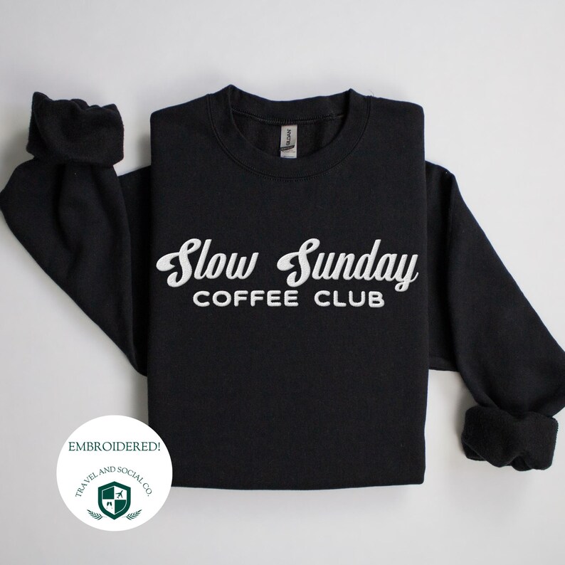 May include: Black sweatshirt with white embroidered text that reads "Slow Sunday Coffee Club". The sweatshirt is folded and displayed on a white background. A white circle with green text and a logo reads "Embroidered! Travel and Social Co."