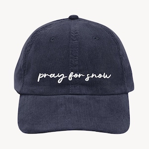 May include: Navy blue corduroy baseball cap with the phrase "pray for snow" in white script across the front. The cap has a curved brim and a structured crown, perfect for casual wear.