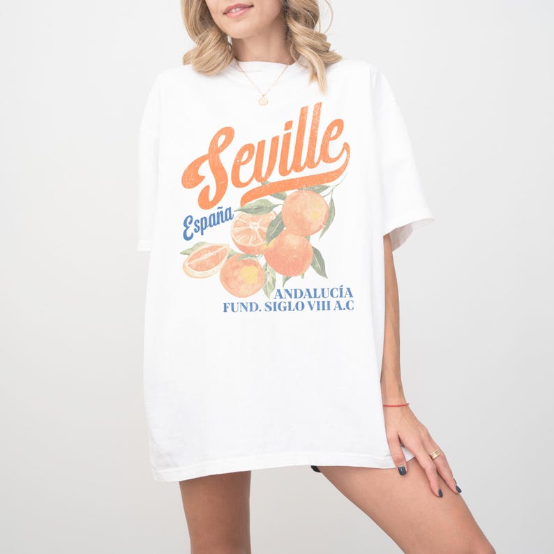 Pu&ograve; includere: Maglietta bianca con una stampa grafica di arance e il testo "Seville, Espa&ntilde;a, Andaluc&iacute;a, Fund. Siglo VIII A.C."