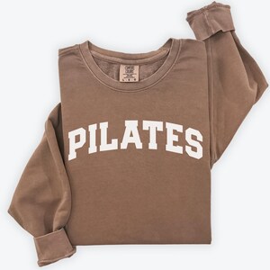 May include: A brown sweatshirt with the word "PILATES" in large, white, block letters across the front. The sweatshirt has a crew neck and long sleeves with cuffed wrists. The garment is folded neatly on a white surface.