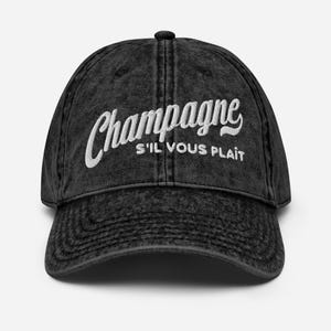 May include: A black baseball cap with the word "Champagne" embroidered in white script. Below the word "Champagne" is the phrase "S'IL VOUS PLAIT" in white lettering. The cap has a curved brim and a button on top.
