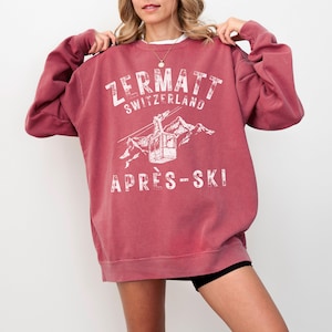 May include: A maroon sweatshirt with a white graphic print of a ski lift in the mountains. The text on the sweatshirt reads "Zermatt Switzerland Apr&egrave;s-Ski".