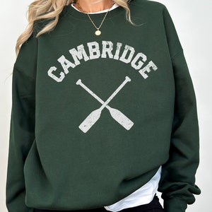 May include: A dark green sweatshirt with a white distressed graphic of crossed oars and the word "CAMBRIDGE" in a white arched font.