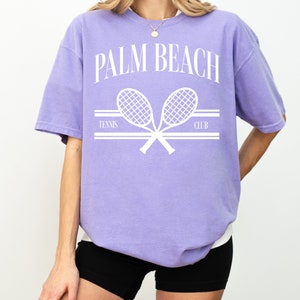 May include: A lavender t-shirt with a white graphic of two tennis rackets crossed over each other. The text "PALM BEACH TENNIS CLUB" is printed above the rackets.