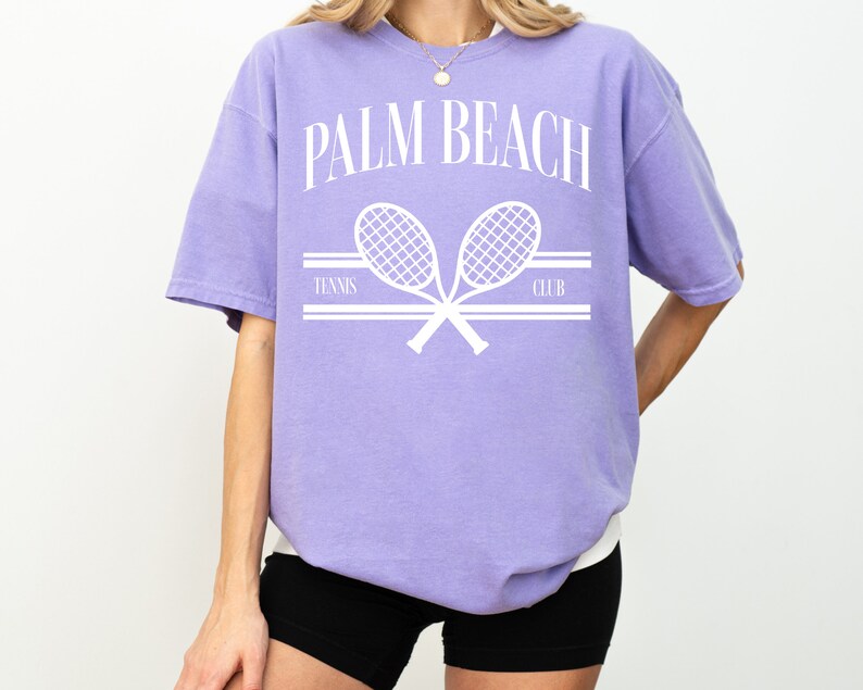 May include: A lavender t-shirt with a white graphic of two tennis rackets crossed over each other. The text "PALM BEACH TENNIS CLUB" is printed above the rackets.