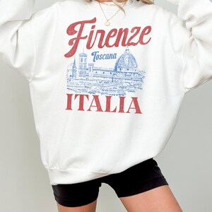 May include: White sweatshirt with a red and blue graphic. The graphic features the word "Firenze" in red script above a blue illustration of a building and the word "ITALIA" in red block letters.