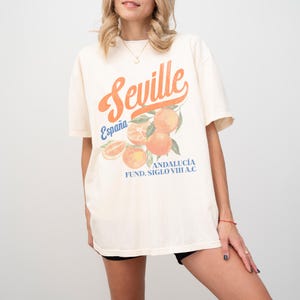 Pu&ograve; includere: Una t-shirt color crema con una stampa grafica di arance e il testo "Sevilla, Espa&ntilde;a, Andaluc&iacute;a, Fund. Siglo VIII A.C."