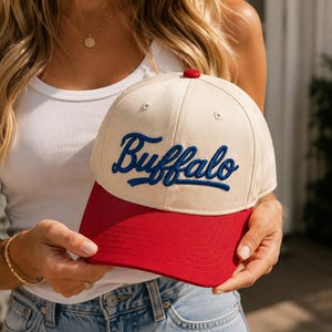 Può includere: Un cappellino da baseball con corona color crema e visiera rossa a tinta unita. La parola Buffalo è ricamata in corsivo blu sui pannelli frontali. Il cappellino presenta un piccolo bottone rosso sulla sommità e cuciture sulla visiera.