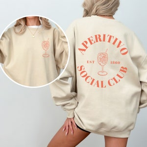 May include: A beige sweatshirt with the text "Aperitivo Social Club Est 1860" in orange on the back. The front of the sweatshirt has a small orange illustration of a cocktail glass.