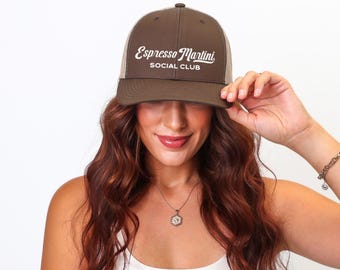 Puede incluir: Gorra de camionero marrón y beige con el texto blanco "Espresso Martini SOCIAL CLUB". La gorra está siendo sostenida. La persona tiene el pelo largo, ondulado y castaño.