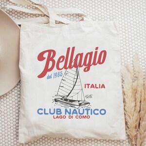 Puede incluir: Bolsa de tela blanca con un dise&ntilde;o gr&aacute;fico rojo y azul. El dise&ntilde;o presenta un velero, el texto "Bellagio dal 1985 ITALIA", y el texto "CLUB NAUTICO LAGO DI COMO".