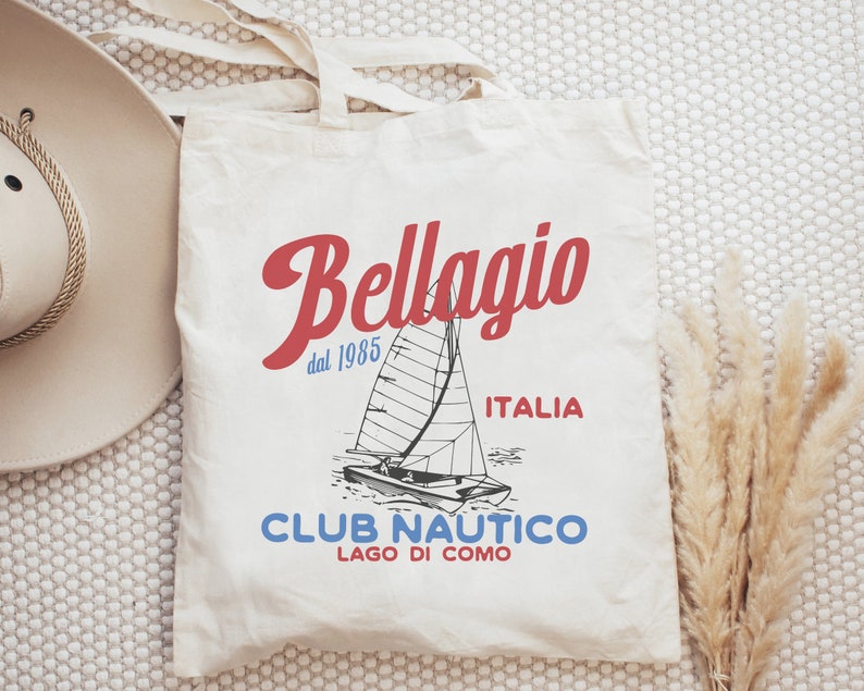 Puede incluir: Bolsa de tela blanca con un dise&ntilde;o gr&aacute;fico rojo y azul. El dise&ntilde;o presenta un velero, el texto "Bellagio dal 1985 ITALIA", y el texto "CLUB NAUTICO LAGO DI COMO".
