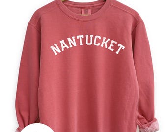 May include: A red-orange crewneck sweatshirt with the word "NANTUCKET" in white block letters across the chest. The sweatshirt has a relaxed fit, long sleeves, and a ribbed neckline, cuffs, and hem. The sleeves are rolled up. A logo is visible on the bottom left.