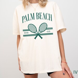May include: An oversized white t-shirt featuring a green graphic of two tennis rackets crossed over each other. The text "PALM BEACH TENNIS CLUB" is printed above the rackets. The shirt is labelled "IVORY" in the bottom right corner.