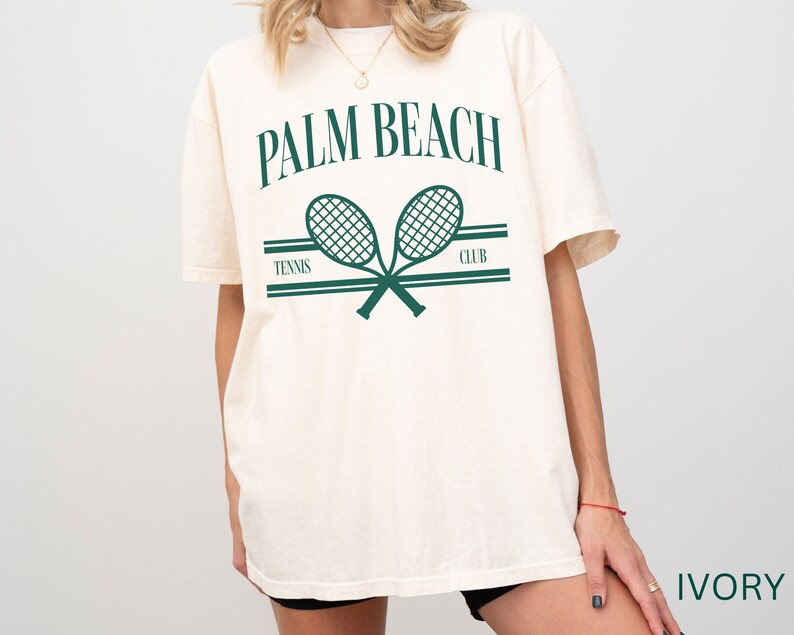 May include: An oversized white t-shirt featuring a green graphic of two tennis rackets crossed over each other. The text "PALM BEACH TENNIS CLUB" is printed above the rackets. The shirt is labelled "IVORY" in the bottom right corner.