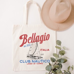 Puede incluir: Una bolsa de lona blanca con un dise&ntilde;o gr&aacute;fico rojo y azul que presenta un velero y el texto "Bellagio dal 1985 ITALIA CLUB NAUTICO LAGO DI COMO".