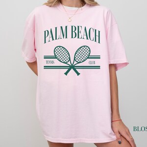 May include: Pink t-shirt featuring a graphic of two tennis rackets crossed over each other. The text "PALM BEACH" is above the rackets and "TENNIS CLUB" is below the rackets.