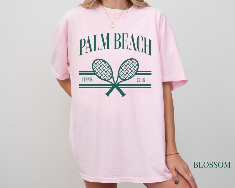 May include: Pink t-shirt featuring a graphic of two tennis rackets crossed over each other. The text "PALM BEACH" is above the rackets and "TENNIS CLUB" is below the rackets.