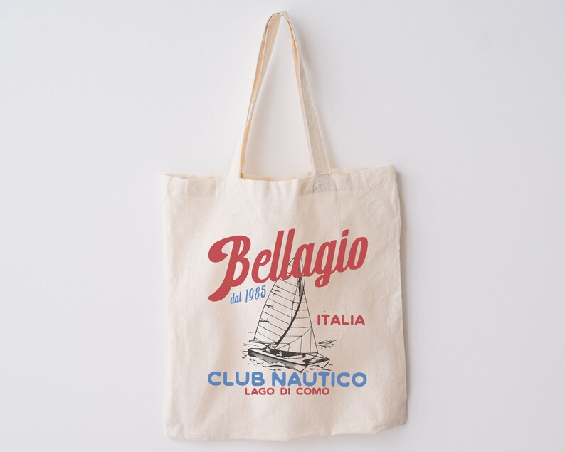 Puede incluir: Una bolsa de lona beige con un dise&ntilde;o gr&aacute;fico rojo y azul. El gr&aacute;fico presenta un velero y el texto "Bellagio dal 1985 ITALIA CLUB NAUTICO LAGO DI COMO".