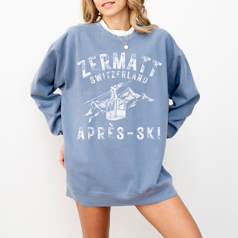 May include: A light blue sweatshirt with a white graphic print of a ski lift in the mountains. The text "Zermatt Switzerland" and "Apr&egrave;s-Ski" are printed on the sweatshirt.