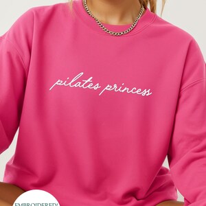 May include: A bright pink sweatshirt with the words "pilates princess" in white script. The sweatshirt has a crew neck and long sleeves. A gold chain necklace is visible at the neckline. The bottom left corner has a circular logo with text.