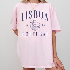 Puede incluir: Camiseta rosa de gran tama&ntilde;o con un dise&ntilde;o gr&aacute;fico azul que presenta una torre, agua y el texto "LISBOA EST 1147 PORTUGAL".