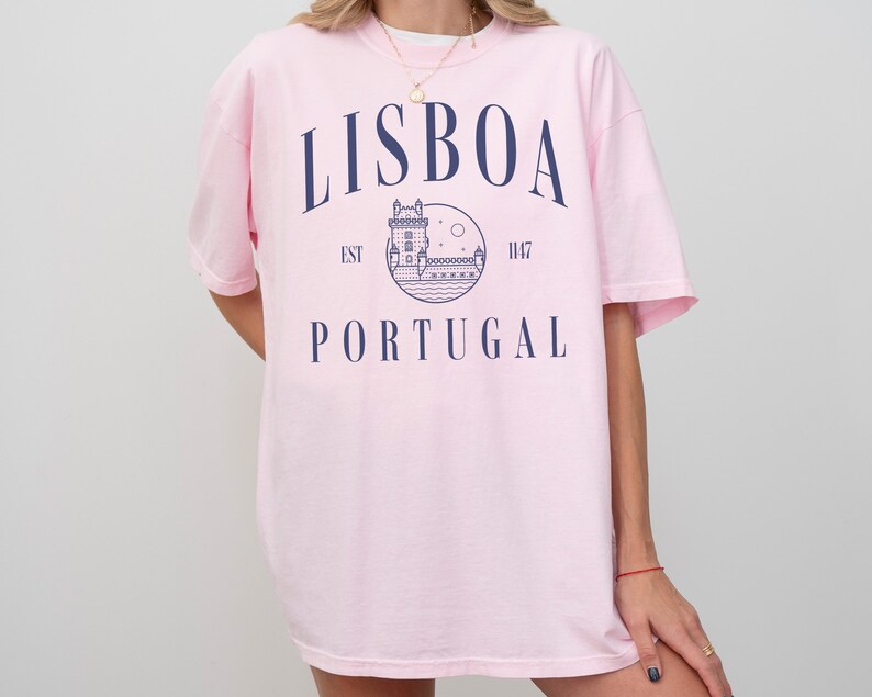 Puede incluir: Camiseta rosa de gran tama&ntilde;o con un dise&ntilde;o gr&aacute;fico azul que presenta una torre, agua y el texto "LISBOA EST 1147 PORTUGAL".