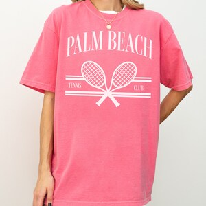 May include: A pink t-shirt with a white graphic of two crossed tennis rackets and the text "PALM BEACH TENNIS CLUB".