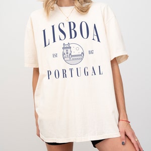 Puede incluir: Una camiseta de color crema con un gr&aacute;fico azul de una torre y el texto "LISBOA EST 1147 PORTUGAL" impreso en la parte delantera.