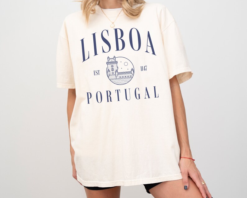 Puede incluir: Una camiseta de color crema con un gr&aacute;fico azul de una torre y el texto "LISBOA EST 1147 PORTUGAL" impreso en la parte delantera.