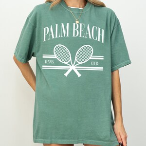 May include: A green t-shirt with a white graphic of two tennis rackets crossed over each other. The text "PALM BEACH" is above the rackets and the text "TENNIS CLUB" is below the rackets.