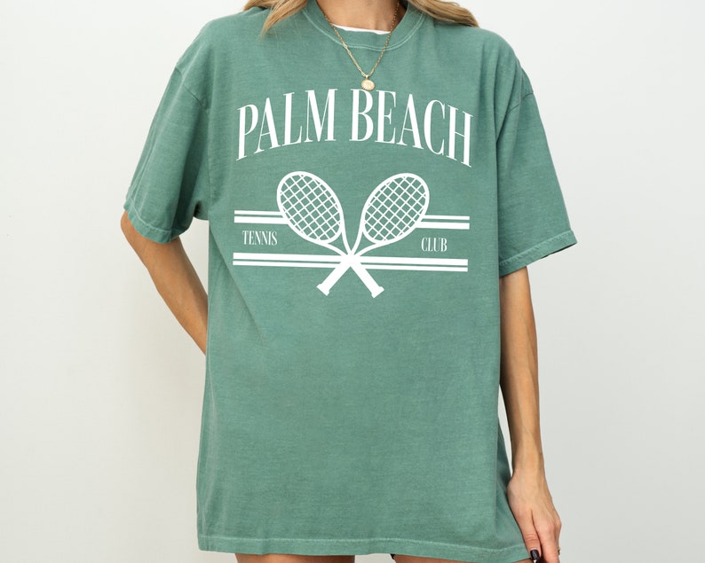 May include: A green t-shirt with a white graphic of two tennis rackets crossed over each other. The text "PALM BEACH" is above the rackets and the text "TENNIS CLUB" is below the rackets.