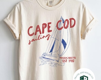 May include: A cream-colored short-sleeve t-shirt featuring a graphic of a sailboat on the water. The text reads CAPE COD sailing MASSACHUSETTS EST 1980 in red and blue ink. The shirt is displayed on a metal hanger against a plain white wall.