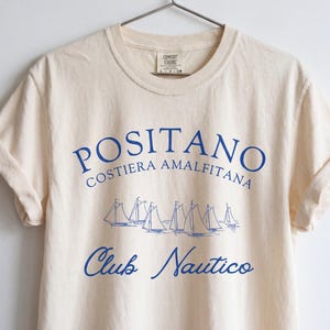 May include: A cream-colored short-sleeve t-shirt featuring blue text that reads POSITANO COSTIERA AMALFITANA and Club Nautico, with a graphic illustration of several sailboats in the center. The shirt has a crew neckline and is on a hanger.