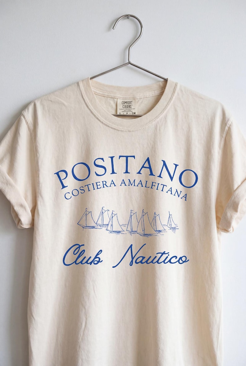 May include: A cream-colored short-sleeve t-shirt featuring blue text that reads POSITANO COSTIERA AMALFITANA and Club Nautico, with a graphic illustration of several sailboats in the center. The shirt has a crew neckline and is on a hanger.