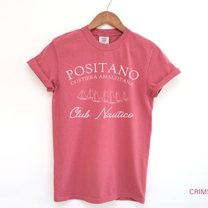May include: A crimson short-sleeve t-shirt featuring white screen-printed text that reads POSITANO COSTIERA AMALFITANA and Club Nautico, with a graphic illustration of several sailboats positioned between the two lines of text.