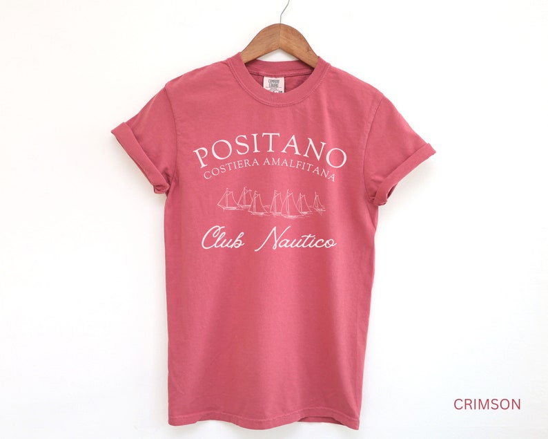 May include: A crimson short-sleeve t-shirt featuring white screen-printed text that reads POSITANO COSTIERA AMALFITANA and Club Nautico, with a graphic illustration of several sailboats positioned between the two lines of text.