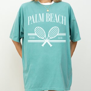 May include: An oversized teal blue t-shirt featuring a white graphic of two tennis rackets crossed over each other. The text "PALM BEACH TENNIS CLUB" is printed above the rackets.