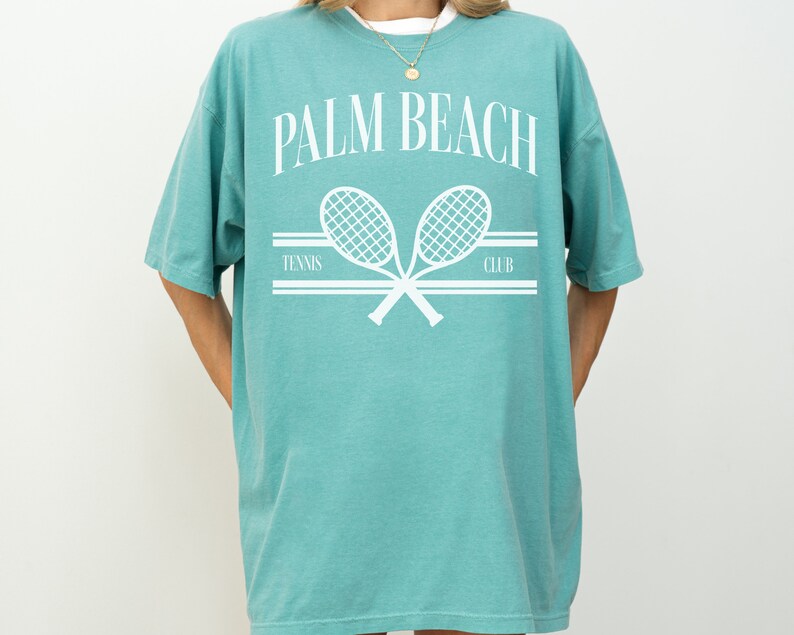 May include: An oversized teal blue t-shirt featuring a white graphic of two tennis rackets crossed over each other. The text "PALM BEACH TENNIS CLUB" is printed above the rackets.