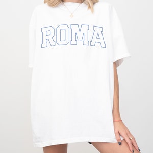 May include: White oversized t-shirt with a blue outline of the word "ROMA" printed on the front.