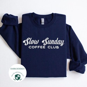 May include: Navy blue sweatshirt with white embroidered text that reads "Slow Sunday Coffee Club". The sweatshirt is folded in half and the text is centered on the front of the sweatshirt. The sweatshirt is embroidered with a small green logo that reads "Travel and Social Co."